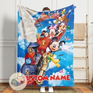 May include: A blue blanket featuring a colorful illustration of Mickey Mouse, Minnie Mouse, Donald Duck, and other Disney characters on a train and in a hot air balloon. The blanket has the text "CUSTOM NAME" in red.