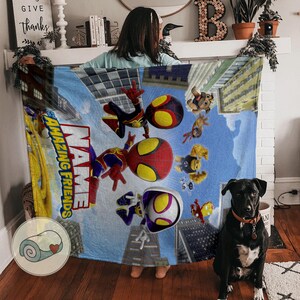 May include: A blue blanket featuring cartoon superheroes and the text "NAME AMAZING FRIENDS" in white and yellow. The blanket has a cityscape background with tall buildings and a rolled-up blanket graphic in the corner.
