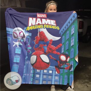 May include: A blue Marvel blanket featuring Spider-Man and his Amazing Friends characters in a cityscape design. The blanket has a large logo with the words "NAME" and "AMAZING FRIENDS". The blanket is held up by a person.