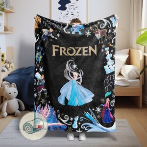 Op de afbeelding: Zwarte fleece deken met personages en afbeeldingen uit de film "Frozen". De deken heeft het woord "FROZEN" in goud, met illustraties van Elsa, Anna, Olaf en andere personages. De deken is opgerold in de linkerbenedenhoek.