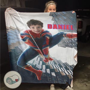 Op de afbeelding: Een gepersonaliseerde deken met een jonge jongen verkleed als Spider-Man, met de naam "DANIEL" in rode en blauwe letters. De deken heeft een stadsgezicht achtergrond en een klein logo van een opgerolde deken met een hart.