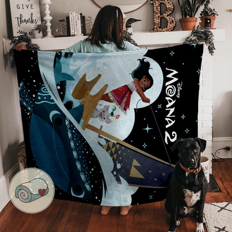 May include: A black blanket featuring a colorful illustration of Moana from Disney's Moana 2. The design includes Moana, a boat, and other characters against a night sky background. The blanket is held up by a person.