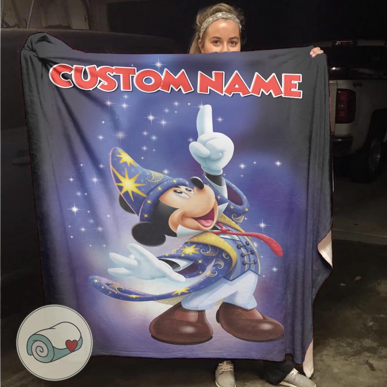 Op de afbeelding: Een donkerblauwe deken met een cartoonafbeelding van Mickey Mouse verkleed als tovenaar, met de tekst "CUSTOM NAME" in het rood. De deken heeft een sterrenachtergrond en een logo van een opgerolde deken.