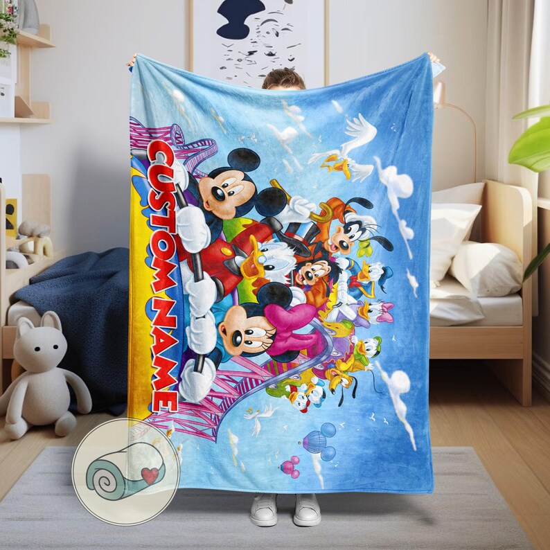 May include: A blue blanket featuring a colorful illustration of Mickey Mouse, Minnie Mouse, Donald Duck, Goofy, and other Disney characters. The blanket has the text "CUSTOM NAME" in a vertical design. The blanket is held up by a person.