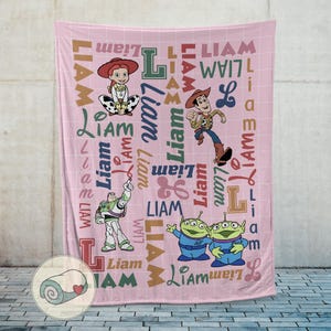 May include: Pink fleece blanket featuring Toy Story characters Jessie, Woody, Buzz Lightyear, and the Little Green Men. The name "Liam" is printed in various fonts and colors across the blanket. A rolled-up blanket with a heart is in the corner.