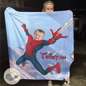 Op de afbeelding: Een lichtblauwe deken met een gepersonaliseerde afbeelding van een kind verkleed als Spider-Man. Het kind wordt afgebeeld in een dynamische pose met een web, tegen een stadsgezicht. De naam "Tristan" is in het rood geschreven.
