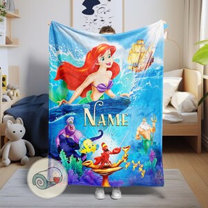 May include: A blue blanket featuring Ariel from The Little Mermaid, with a red-haired mermaid, a ship, and other characters. The word "Name" is written on the blanket. The blanket is displayed in a bedroom setting.