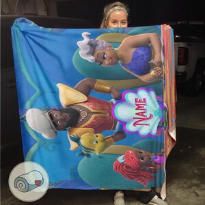 May include: A blue blanket with cartoon characters from a movie. The blanket has a seashell-shaped area with the word "NAME" on it. The characters include a mermaid, a king, and a yellow fish.