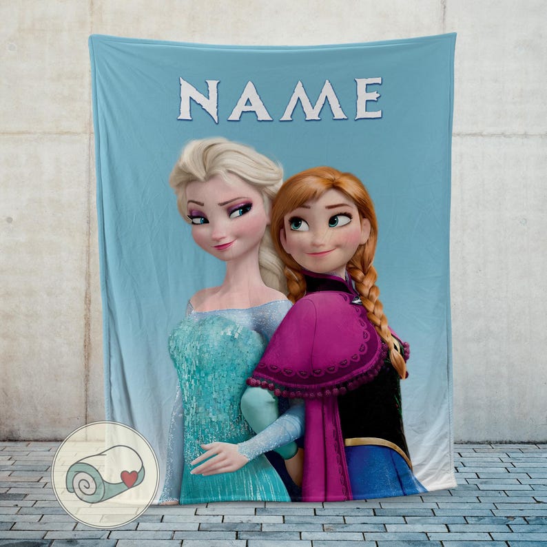 Puede incluir: Una manta azul claro con los personajes Elsa y Anna de la pel&iacute;cula Frozen. La manta tiene la palabra "NAME" en letras blancas en la parte superior. Elsa lleva un vestido azul y Anna una capa morada.