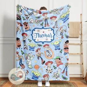 May include: A light blue blanket featuring various cartoon characters, including Woody, Buzz Lightyear, and others. The blanket has a repeating pattern of the characters and includes the name "Thomas" in a decorative frame. The blanket is made of a soft material.