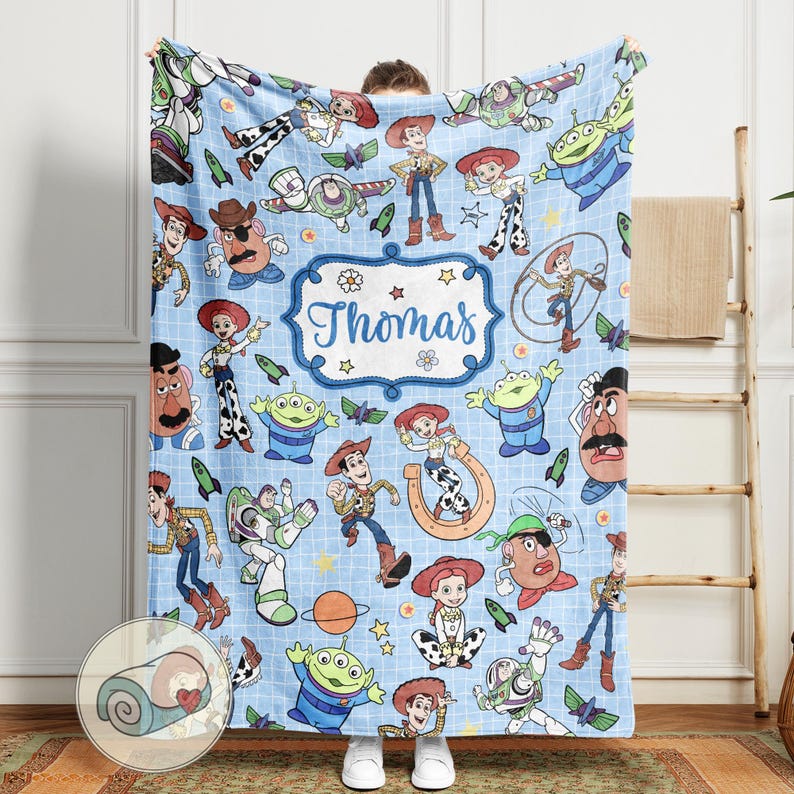 May include: A light blue blanket featuring various cartoon characters, including Woody, Buzz Lightyear, and others. The blanket has a repeating pattern of the characters and includes the name "Thomas" in a decorative frame. The blanket is made of a soft material.