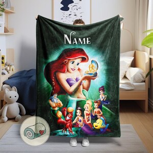 May include: A dark green plush blanket featuring a colorful illustration of Ariel from The Little Mermaid. The design includes Ariel, Flounder, Sebastian, and other characters. The word "Name" is written in white script at the top.