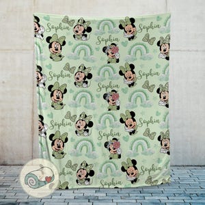 K&ouml;nnte beinhalten: Eine hellgr&uuml;ne Decke mit einem sich wiederholenden Muster von Minnie Mouse, Schleifen, Regenbogen, Wolken und Herzen. Der Name "Sophia" ist in Schreibschrift geschrieben. Die Decke besteht aus einem weichen Material.