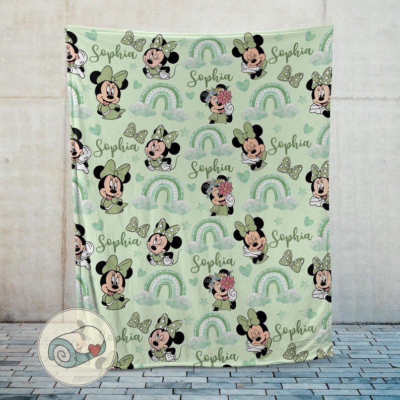 K&ouml;nnte beinhalten: Eine hellgr&uuml;ne Decke mit einem sich wiederholenden Muster von Minnie Mouse, Schleifen, Regenbogen, Wolken und Herzen. Der Name "Sophia" ist in Schreibschrift geschrieben. Die Decke besteht aus einem weichen Material.