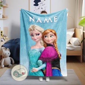 Puede incluir: Una manta azul claro con Elsa y Anna de Frozen, con la palabra "NAME" en letras blancas en la parte superior. La manta tiene una ilustraci&oacute;n de dibujos animados de los personajes y la sostiene una persona.