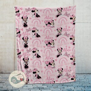K&ouml;nnte beinhalten: Rosa Decke mit einem sich wiederholenden Muster von Minnie Mouse, Schleifen, Regenbogen, Sternen, Wolken und dem Namen "Alice". Die Decke besteht aus einem weichen Material und ist f&uuml;r Kinder geeignet.