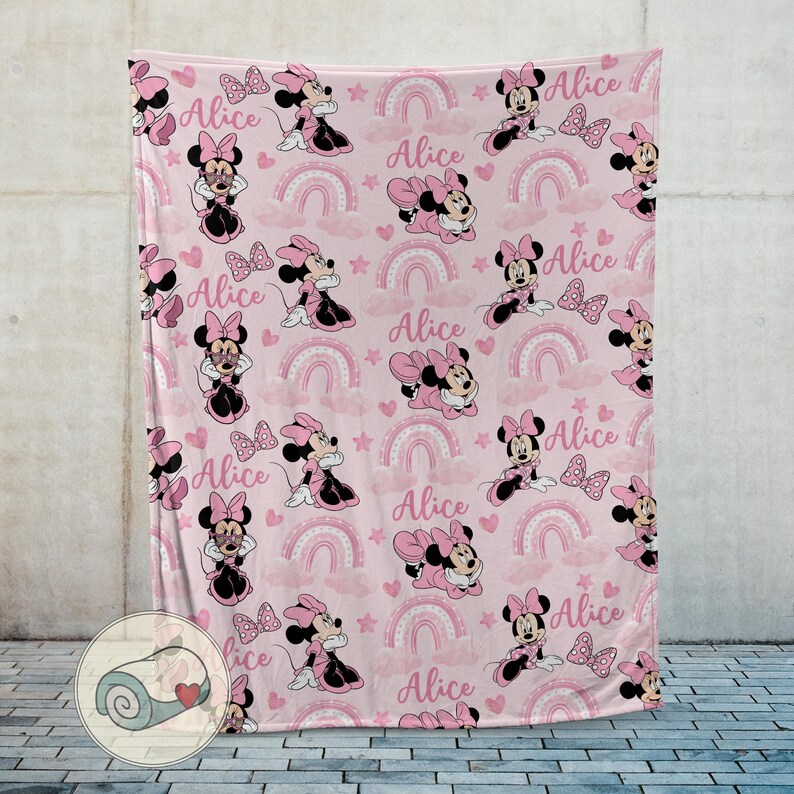 K&ouml;nnte beinhalten: Rosa Decke mit einem sich wiederholenden Muster von Minnie Mouse, Schleifen, Regenbogen, Sternen, Wolken und dem Namen "Alice". Die Decke besteht aus einem weichen Material und ist f&uuml;r Kinder geeignet.