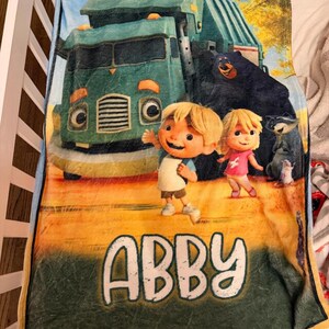 Pu&ograve; includere: Una coperta di peluche con una scena a cartoni animati con un camion verde, due bambini e diversi animali. La coperta ha il nome "ABBY" in grandi lettere bianche in basso. Lo sfondo &egrave; un mix di giallo, verde e marrone.
