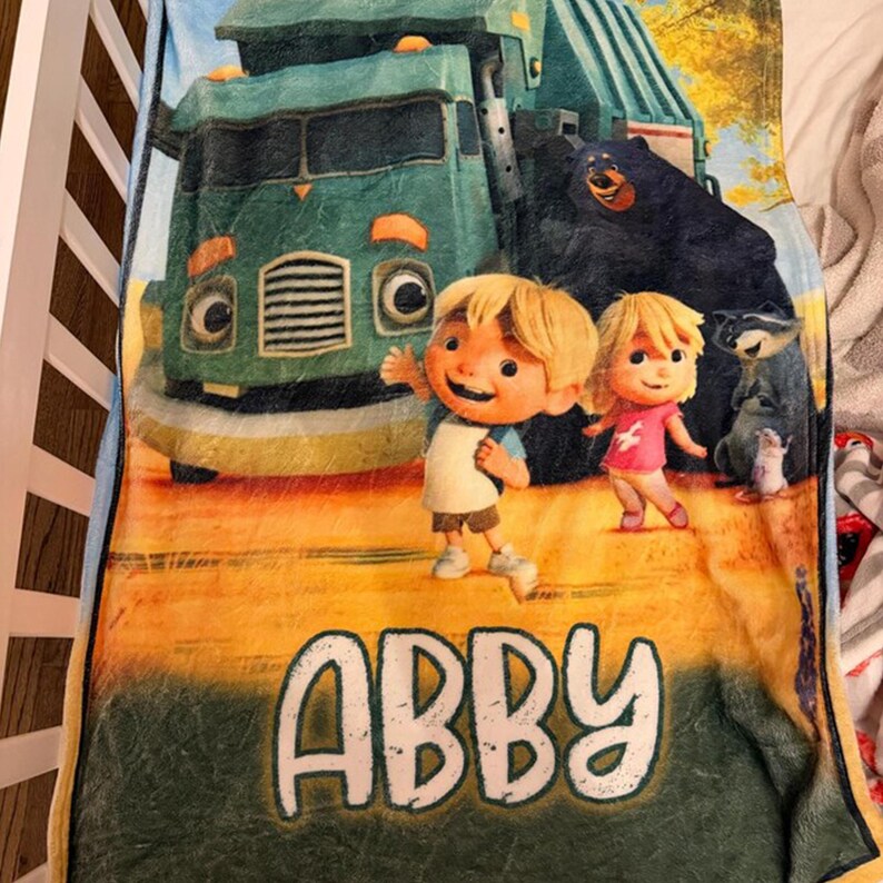 Pu&ograve; includere: Una coperta di peluche con una scena a cartoni animati con un camion verde, due bambini e diversi animali. La coperta ha il nome "ABBY" in grandi lettere bianche in basso. Lo sfondo &egrave; un mix di giallo, verde e marrone.