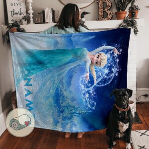 Op de afbeelding: Een blauw-witte deken met een afbeelding van een prinses in een vloeiende jurk en een kasteel. De deken heeft de naam "NAM" erop gedrukt. Een zwarte hond met een bruine halsband zit voor de deken.