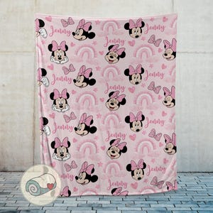 May include: Pink fleece blanket featuring a repeating pattern of Minnie Mouse, bows, rainbows, hearts, and the name "Jenny" in a script font. The blanket is displayed against a neutral wall.