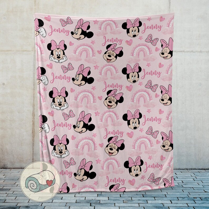 May include: Pink fleece blanket featuring a repeating pattern of Minnie Mouse, bows, rainbows, hearts, and the name "Jenny" in a script font. The blanket is displayed against a neutral wall.