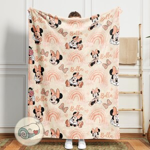 May include: A beige blanket featuring a repeating pattern of Minnie Mouse, bows, and rainbows in shades of pink and peach. The name "Bella" is written in a script font throughout the design. The blanket is soft and cozy.