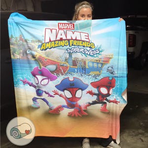 Peut inclure: Une grande couverture rectangulaire avec le logo Marvel et les mots "NAME Amazing Friends Water-Webs". La couverture pr&eacute;sente des images de dessins anim&eacute;s de personnages de Spider-Man en tenue de pirate, un bateau pirate et un requin sur une plage et un fond marin.