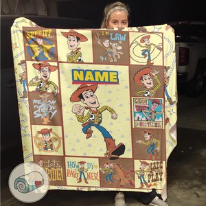 May include: A yellow and brown blanket featuring multiple images of a cartoon cowboy character. The blanket has the word "NAME" in large blue letters and other phrases like "Reach for the Sky" and "Howdy Partner!"