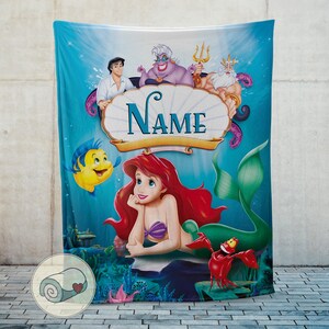 May include: A teal blanket featuring Ariel from The Little Mermaid, with other characters like Flounder, Sebastian, and Prince Eric. The blanket has a personalized name banner and a light blue background.