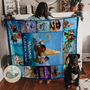 Może przedstawiać: Niebieski koc z motywem Moany z r&oacute;żnymi scenami i postaciami z filmu. Koc ma napis "Moana Custom Name" i inne frazy. Koc jest prezentowany z psem siedzącym w pobliżu.