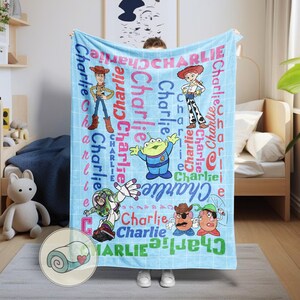 May include: A light blue blanket featuring characters from the Toy Story franchise, including Woody, Buzz Lightyear, and others. The name "Charlie" is printed in various colors across the blanket. The blanket is displayed indoors.