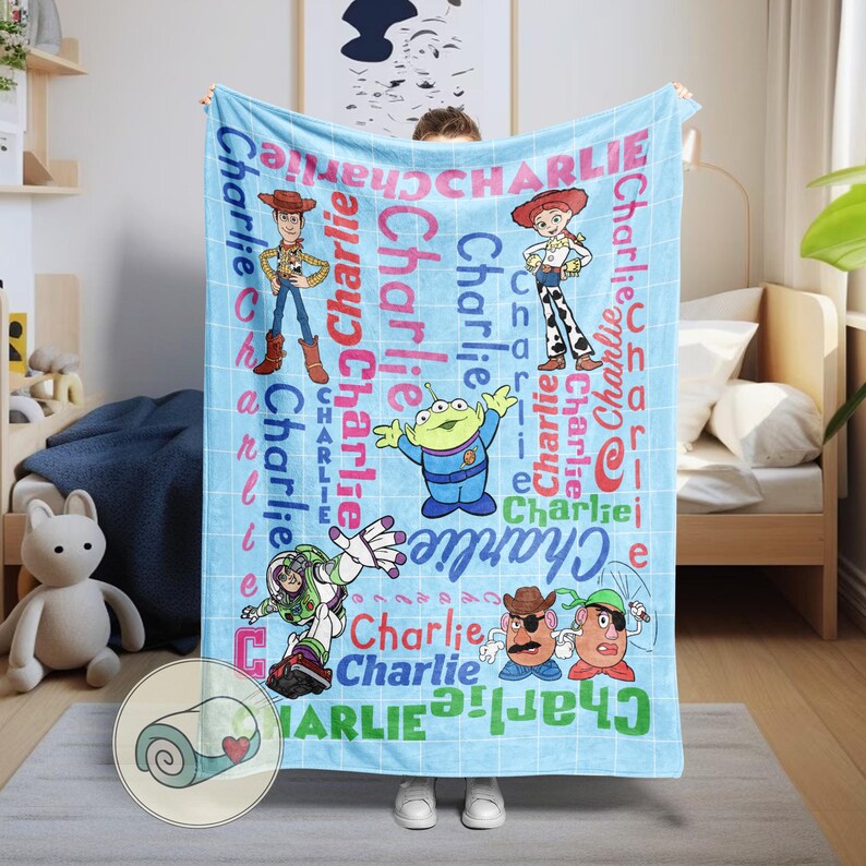 May include: A light blue blanket featuring characters from the Toy Story franchise, including Woody, Buzz Lightyear, and others. The name "Charlie" is printed in various colors across the blanket. The blanket is displayed indoors.