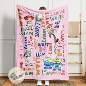 May include: Pink blanket featuring characters from the Toy Story movie, including Jessie, Woody, Buzz Lightyear, and the Little Green Men. The name "Liam" is printed in various fonts across the blanket. The blanket is a soft, cozy item.