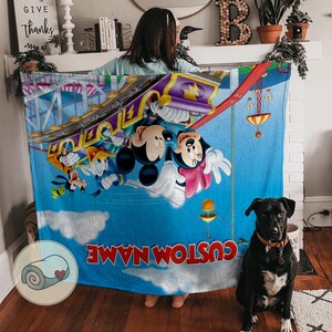 May include: A blue blanket featuring Mickey and Minnie Mouse on a roller coaster, with Goofy and Donald Duck. The blanket has a custom name in red text and a rolled blanket logo.