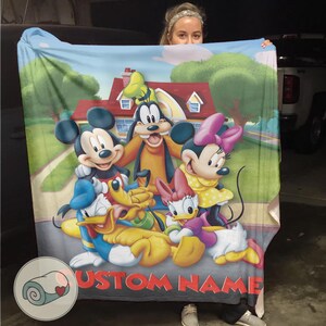 K&ouml;nnte beinhalten: Eine gro&szlig;e, rechteckige Decke mit einer farbenfrohen Illustration von Micky Maus, Minnie Maus, Goofy, Donald Duck, Daisy Duck und Pluto. Die Decke hat einen individuellen Namen, der unten in Rot aufgedruckt ist. Der Hintergrund zeigt ein Haus und eine Stra&szlig;e.