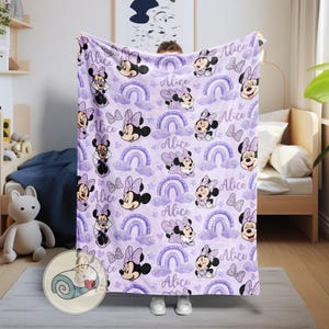 Op de afbeelding: Een lavendelkleurige deken met een herhalend patroon van Minnie Mouse, regenbogen, wolken, sterren en de naam "Alice". De deken heeft een zacht uiterlijk en is waarschijnlijk gemaakt van fleece of een vergelijkbaar materiaal.