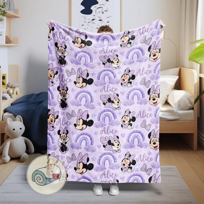 Op de afbeelding: Een lavendelkleurige deken met een herhalend patroon van Minnie Mouse, regenbogen, wolken, sterren en de naam "Alice". De deken heeft een zacht uiterlijk en is waarschijnlijk gemaakt van fleece of een vergelijkbaar materiaal.