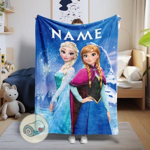 May include: A blue blanket featuring the characters Elsa and Anna from the movie Frozen, with the word "NAME" in white. The blanket has a winter theme with snowflakes and a light blue background. The blanket is being held up.