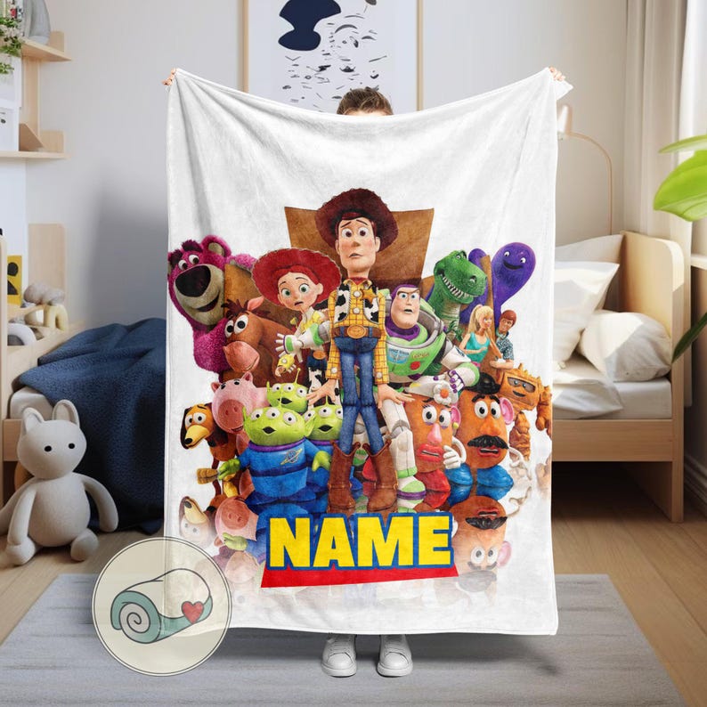 May include: White plush blanket with a vibrant graphic of characters from the Toy Story film. The blanket features Woody, Buzz Lightyear, and other characters. The word "NAME" is printed in yellow on a red stripe.