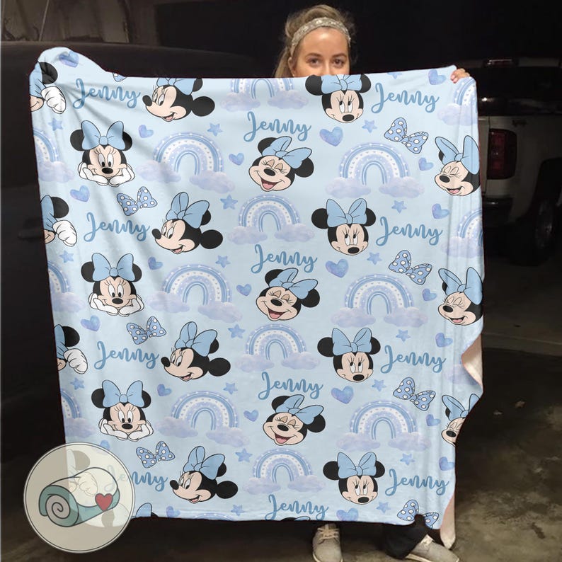 Puede incluir: Una manta azul claro con un estampado de Minnie Mouse, lazos, arco&iacute;ris y corazones. El nombre "Jenny" est&aacute; escrito en cursiva. La manta es suave y acogedora, ideal para la habitaci&oacute;n de un ni&ntilde;o.