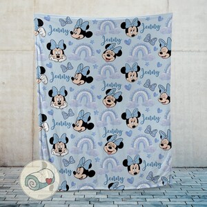 Puede incluir: Una manta azul claro con un estampado de Minnie Mouse, lazos, arco&iacute;ris y corazones. El nombre "Jenny" est&aacute; escrito en el dise&ntilde;o. Una manta suave y acogedora.