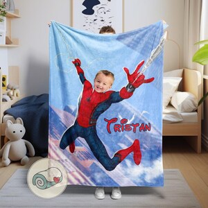 Op de afbeelding: Een blauwe deken met een gepersonaliseerd ontwerp van een kind verkleed als Spider-Man, met de naam "Tristan" in rood schrift. De deken heeft een hemelachtergrond met een web en een bergketen.