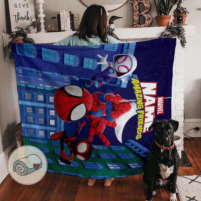 May include: A blue Marvel blanket featuring Spider-Man and Spider-Gwen characters against a cityscape backdrop. The blanket has the words "Amazing Friends" and "Name" printed on it. A black dog with a brown collar sits in front of the blanket.