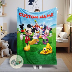 Pu&ograve; includere: Una coperta in pile azzurra con una scena a cartoni animati con Topolino, Minnie, Paperino, Pippo e Pluto. La coperta ha la scritta "CUSTOM NAME" in rosso. I personaggi sono su un campo verde e un cielo blu.