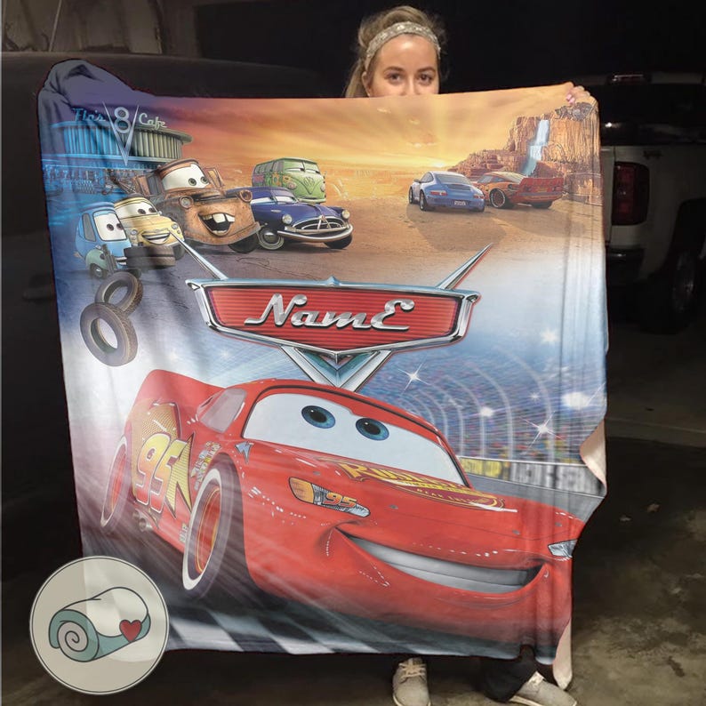 May include: A large, rectangular blanket featuring characters from the animated movie Cars. The blanket showcases Lightning McQueen, Mater, and other vehicles against a desert landscape with the text "Name" on a logo.