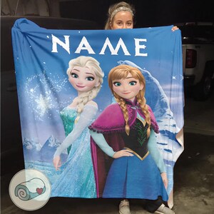 May include: Blue blanket featuring Elsa and Anna from Frozen, with the word "NAME" at the top. The blanket has a wintery background with snowflakes and a mountain. The blanket is being held up by a person.