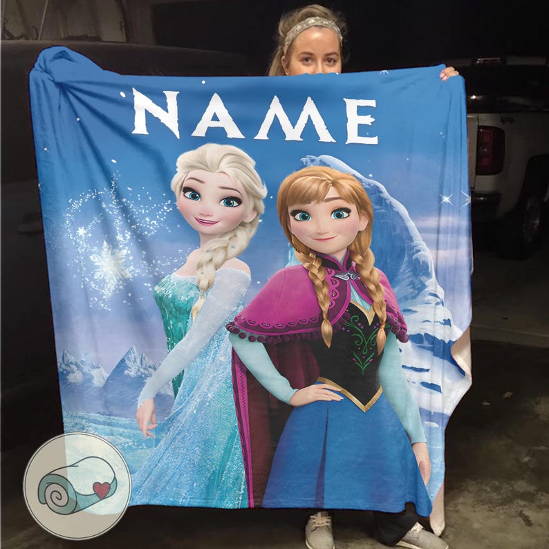 May include: Blue blanket featuring Elsa and Anna from Frozen, with the word "NAME" at the top. The blanket has a wintery background with snowflakes and a mountain. The blanket is being held up by a person.