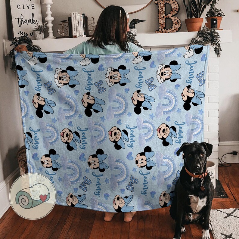 Puede incluir: Una manta azul claro con Minnie Mouse, lazos azules y arco&iacute;ris. El nombre "Jenny" est&aacute; impreso en la manta. Un perro negro con collar marr&oacute;n est&aacute; sentado frente a la manta.