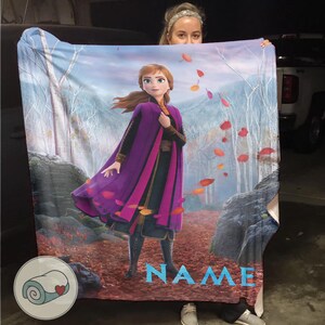 Pode incluir: Uma manta de pel&uacute;cia com uma ilustra&ccedil;&atilde;o vibrante de uma personagem do filme de anima&ccedil;&atilde;o "Frozen". O design apresenta a personagem com uma capa roxa, num cen&aacute;rio de floresta com folhas a cair. A palavra "NAME" est&aacute; impressa em azul.